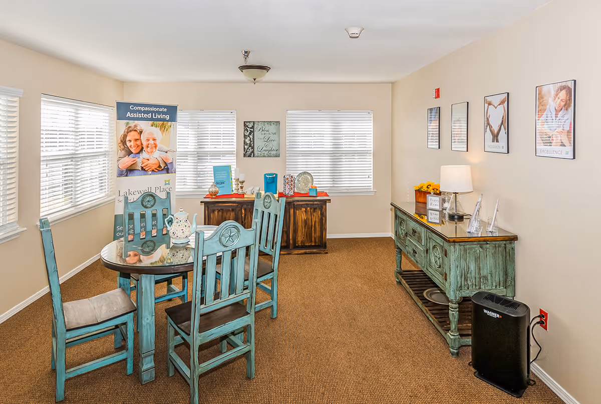 Bright common dining area with a round wooden table and turquoise chairs, a rustic sideboard, and an assisted-living promotional banner.