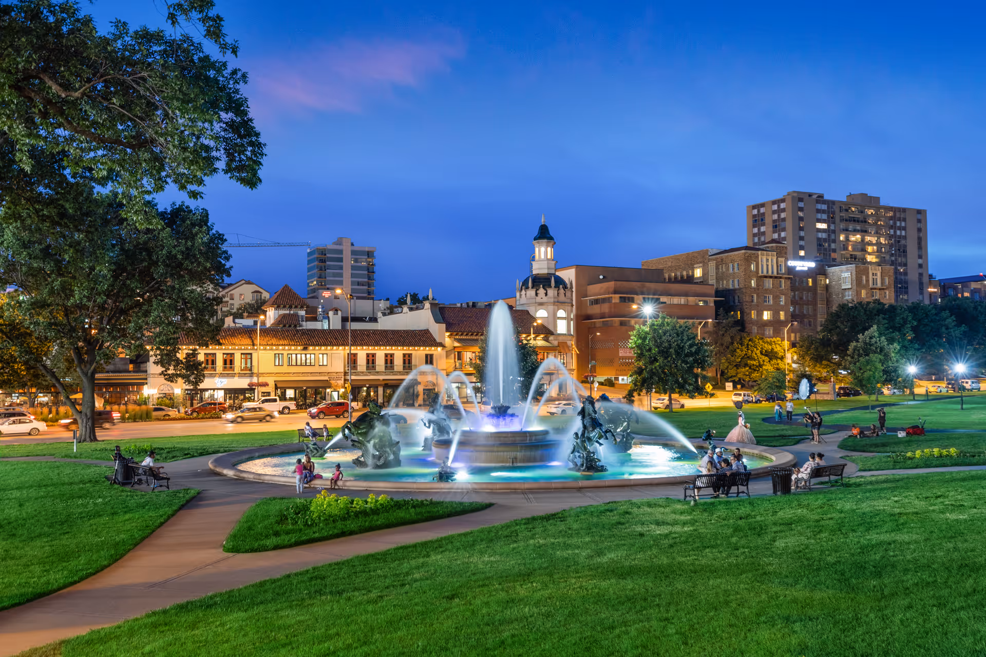 A vibrant city park at dusk featuring a large, illuminated fountain with water jets surrounded by green lawns and walking paths. People are sitting on benches and walking around the park. In the background, there are buildings with lights on, including a tall building and a structure with a small tower.