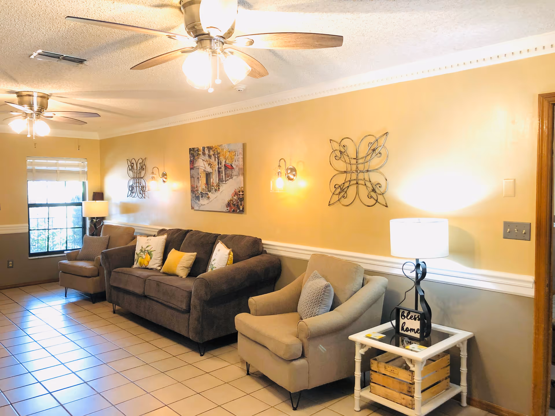 A cozy living room with beige walls and tiled floor featuring a dark gray sofa with decorative pillows, two beige armchairs, a white side table with a lamp and a decorative sign that says 'bless this home'. The room is illuminated by ceiling fans with lights and wall sconces, and there is a window with blinds on the left side.