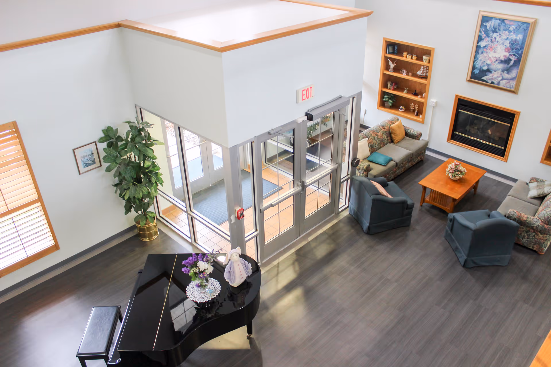 Interior view of a senior living facility lobby with a black grand piano decorated with flowers and a stuffed bunny, a seating area with floral and solid-colored sofas and armchairs around a wooden coffee table, a fireplace with a floral painting above it, large windows, and an exit door.