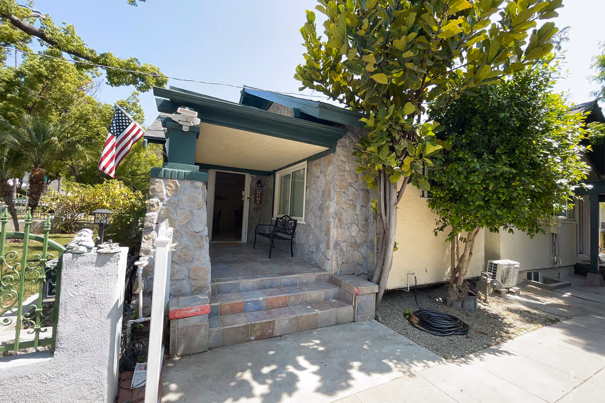 Front entrance of a residential-style building with stone pillars and tiled steps leading up to a covered porch area. There is a black metal bench on the porch, an American flag mounted on the left side, and trees providing shade around the entrance.