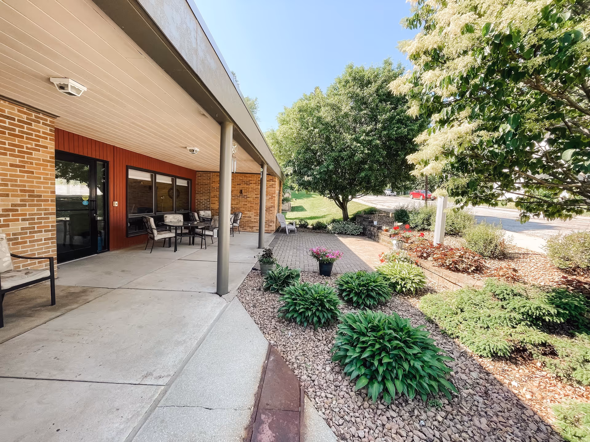 Outdoor patio area at Traditions of Preston with a covered seating area featuring tables and chairs, surrounded by landscaped garden beds with green shrubs, flowering plants, and trees under a clear blue sky.