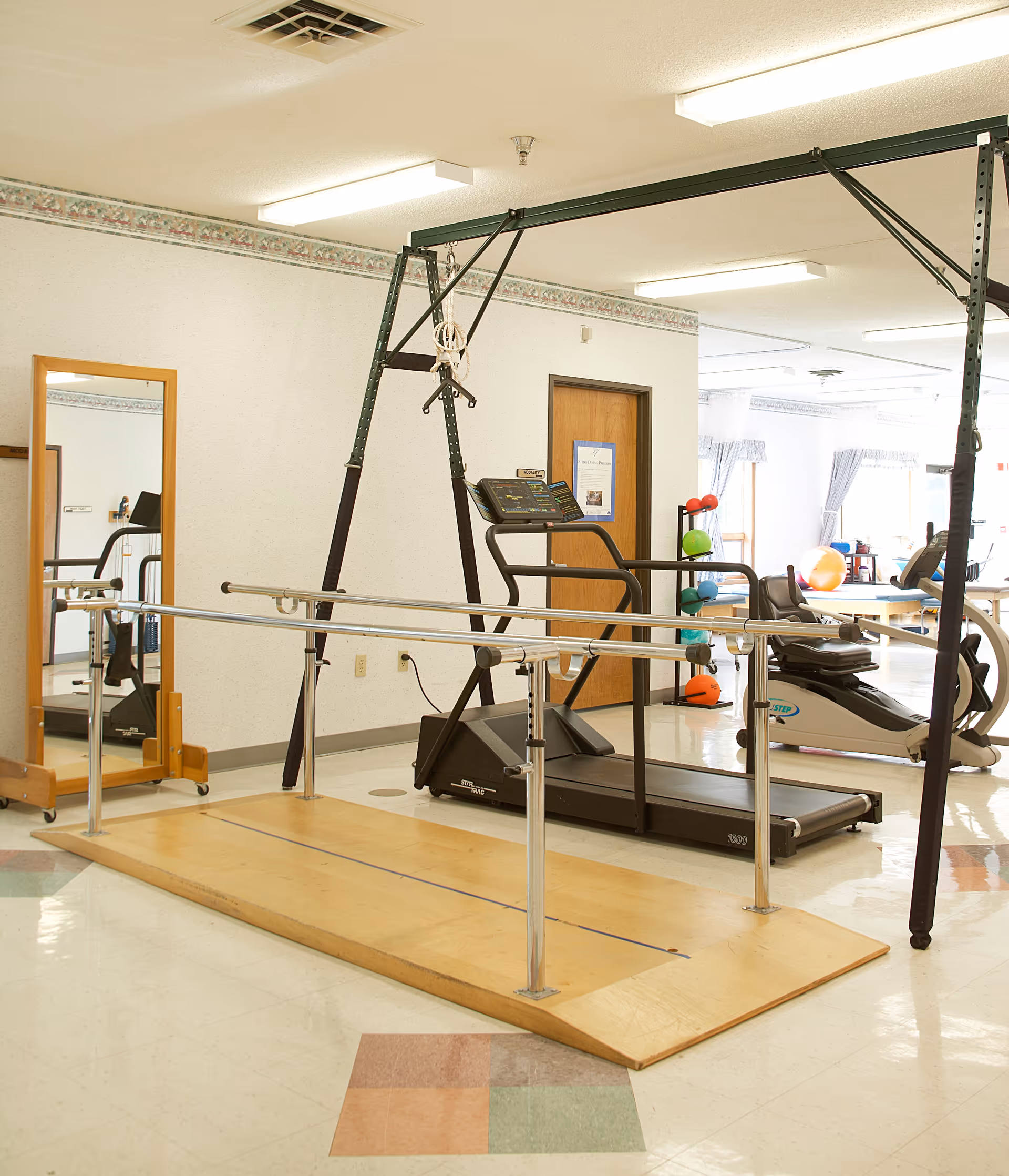 Physical therapy or exercise room with parallel bars on a wooden platform, a treadmill, a recumbent exercise bike, a full-length mirror, and a rack holding colorful exercise balls in a well-lit room with white walls and a door in the background.