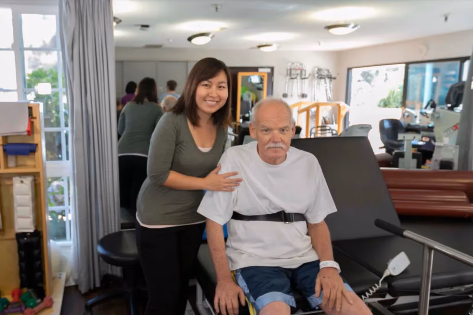 A female caregiver standing next to an elderly man seated on a medical examination table in a rehabilitation or physical therapy room with exercise equipment and walkers in the background.