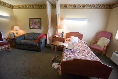 A senior living facility bedroom with a single bed covered in a floral bedspread, a bedside table with a lamp, a green striped couch with a red blanket, a pink armchair with a cushion, a small table with an open book, slippers on the floor, and a floral border wallpaper near the ceiling.