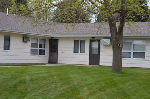 Single-story senior living home exterior with doors, windows, tree, and green lawn.