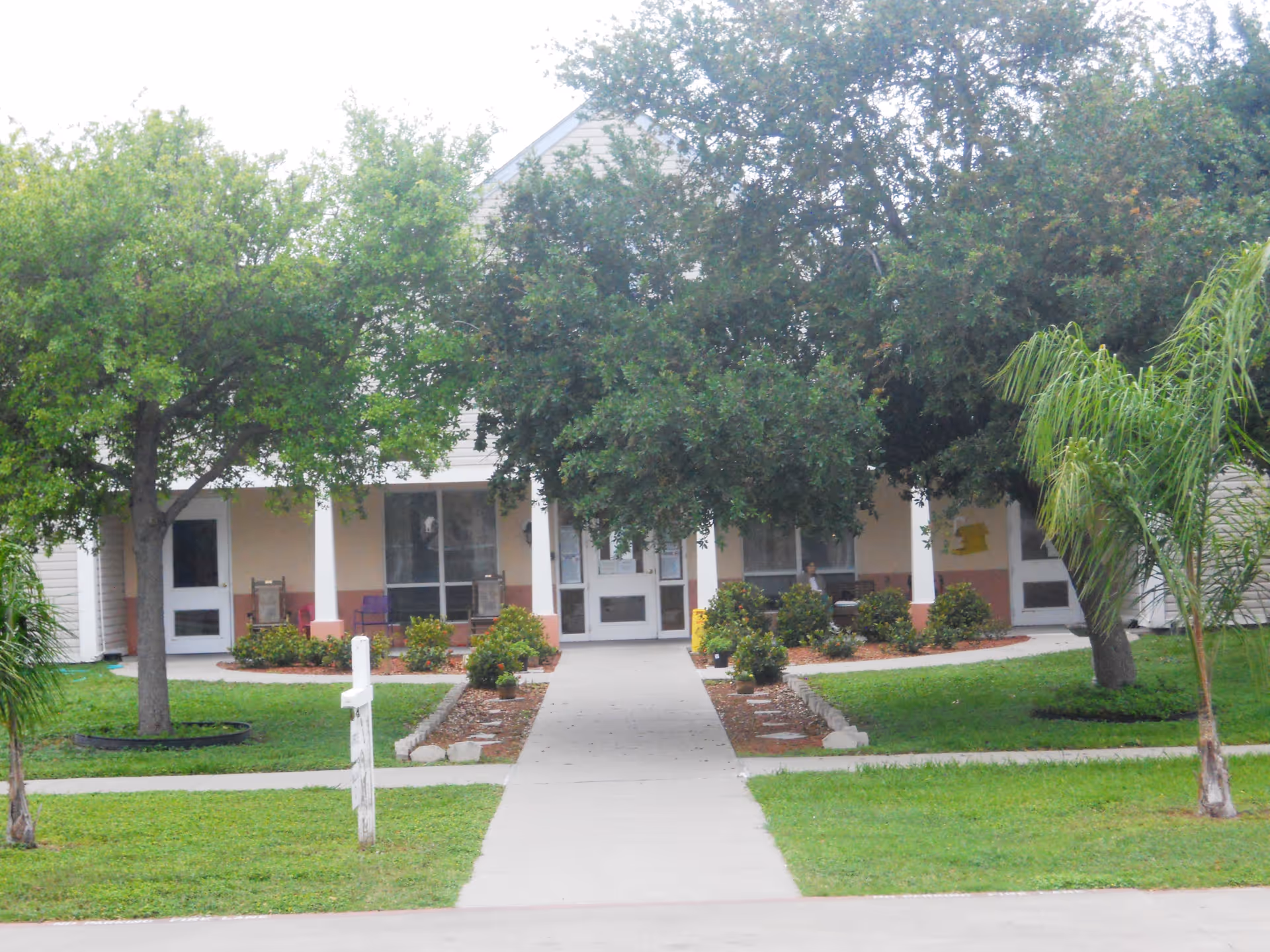 Front entrance of an assisted living building with a paved walkway, porch columns, shrubs, and trees.