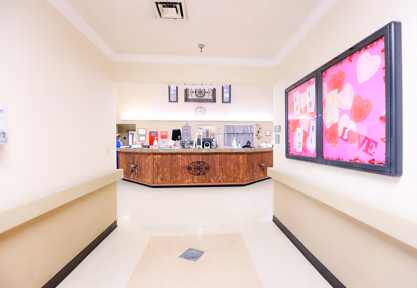 A hallway leading to a reception desk area in a senior living facility. The desk is wooden with decorative elements and various items on top, including hand sanitizer. On the right wall, there is a bulletin board decorated with pink paper and heart shapes, featuring the word 'LOVE'. The walls are painted beige with handrails on both sides of the hallway.