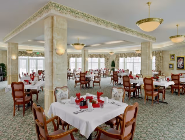 A spacious dining room with multiple tables covered in white tablecloths, each set with red napkins, glasses, and cutlery. The room features large windows allowing natural light, decorative columns, patterned carpet, and elegant light fixtures hanging from the ceiling.