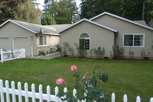 Exterior view of a single-story beige building with white trim, surrounded by a green lawn and a white picket fence. There are pink roses and other plants in front of the building, and tall trees in the background.