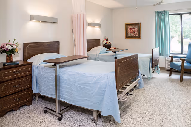 A bright and clean room with two hospital-style beds covered in light blue sheets, each equipped with an overbed table. There is a wooden nightstand with a vase of flowers and a book between the beds. A framed picture hangs on the wall, and a window with teal curtains lets in natural light. Two blue chairs are positioned near the window.