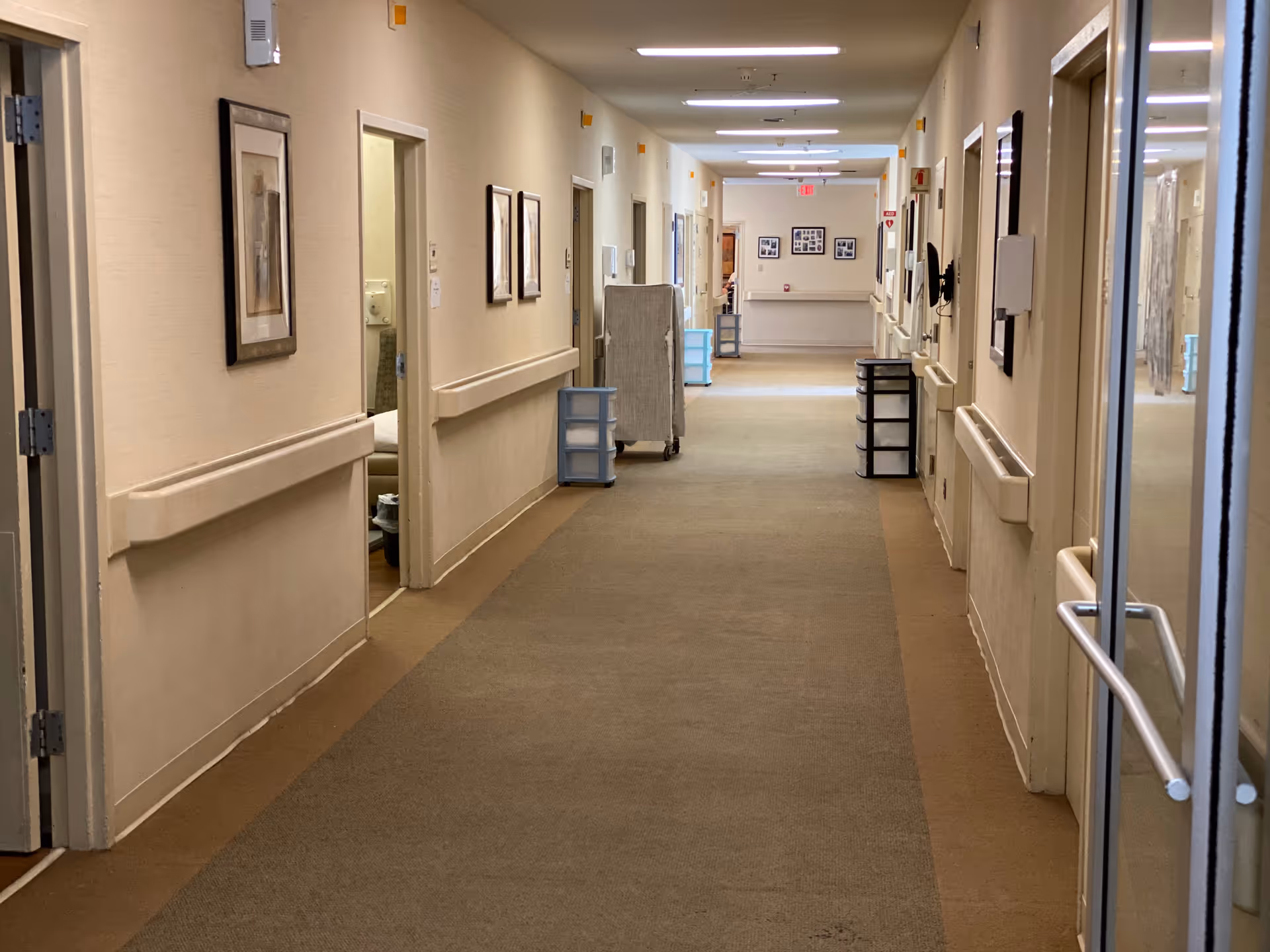 A long carpeted corridor in a senior living facility with handrails, open room doors, framed pictures, and utility carts.