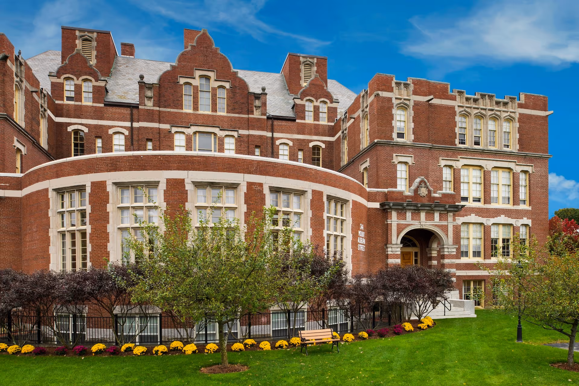 Exterior view of a large, historic brick building with multiple windows and decorative architectural details. The building is surrounded by green grass, trees, and colorful flowers, with a bench near the entrance under a bright blue sky.