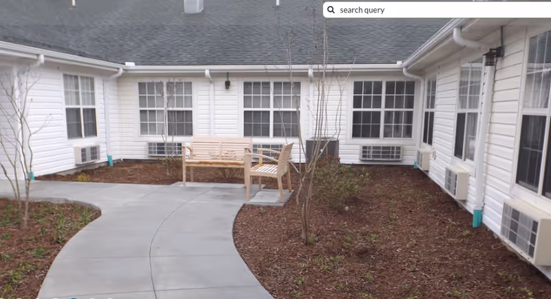 Outdoor courtyard area with a curved concrete walkway, two wooden benches, small trees, and plants surrounded by a white building with multiple windows and air conditioning units.