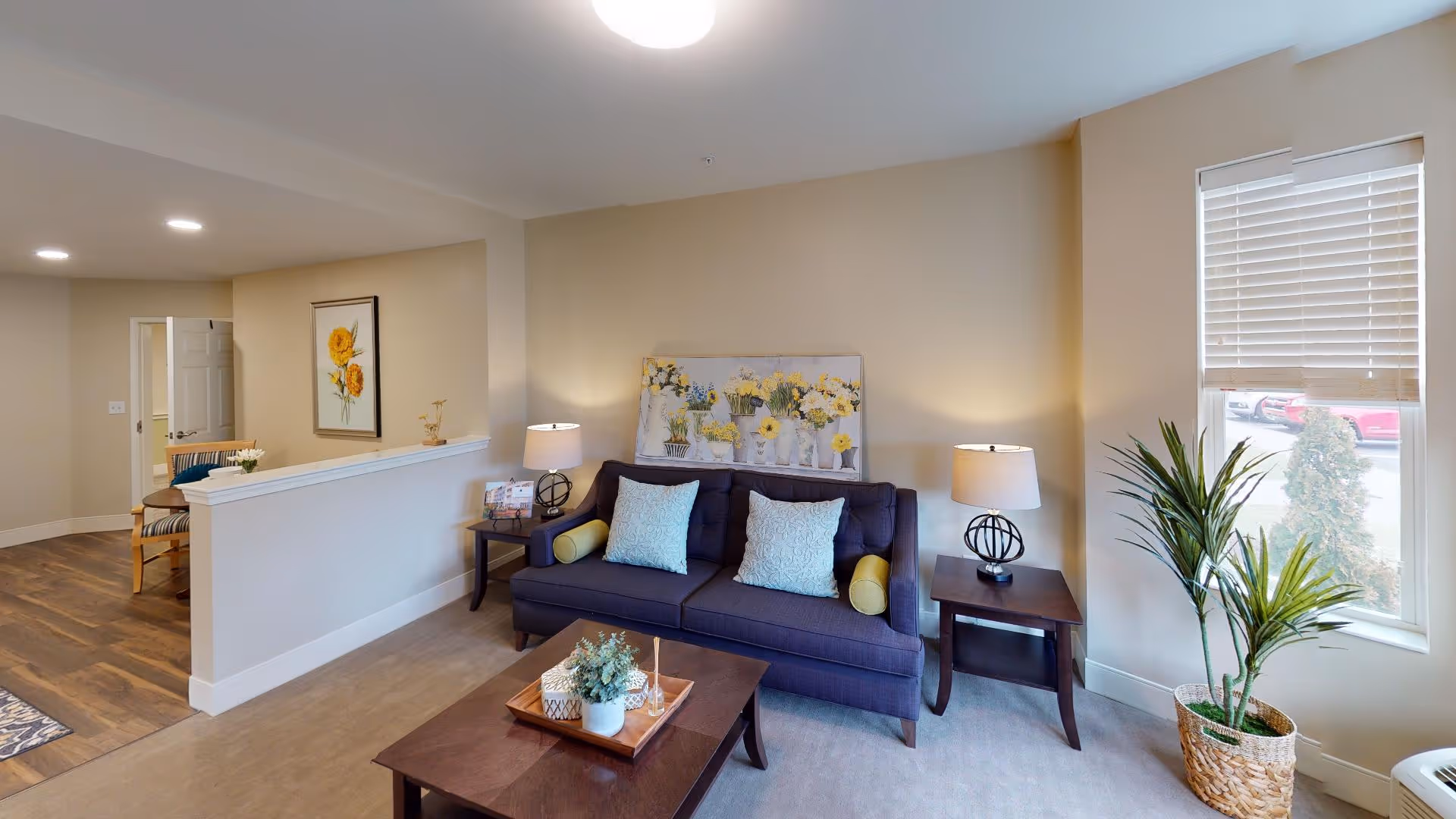 A cozy living room area in a senior living facility featuring a dark blue sofa with light blue and yellow pillows, two side tables each with a lamp, a coffee table with decorative items, a large window with blinds, a potted plant, and a wall painting of yellow flowers. The room has beige walls and carpeted flooring, with an adjacent area showing wooden flooring and a chair.