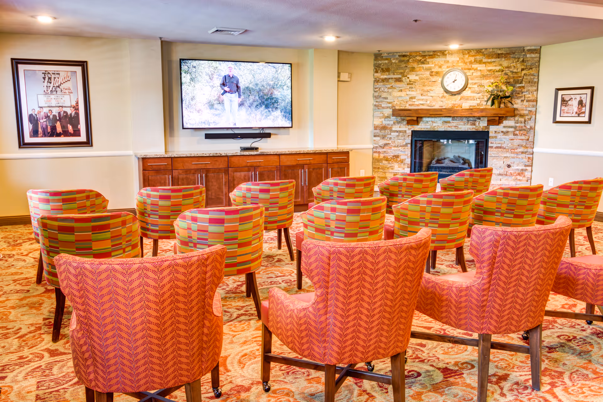 A cozy common area with multiple colorful patterned chairs arranged in rows facing a wall-mounted flat screen TV. The room features a stone fireplace with a clock above it and framed pictures on the walls. The floor is covered with a patterned carpet.