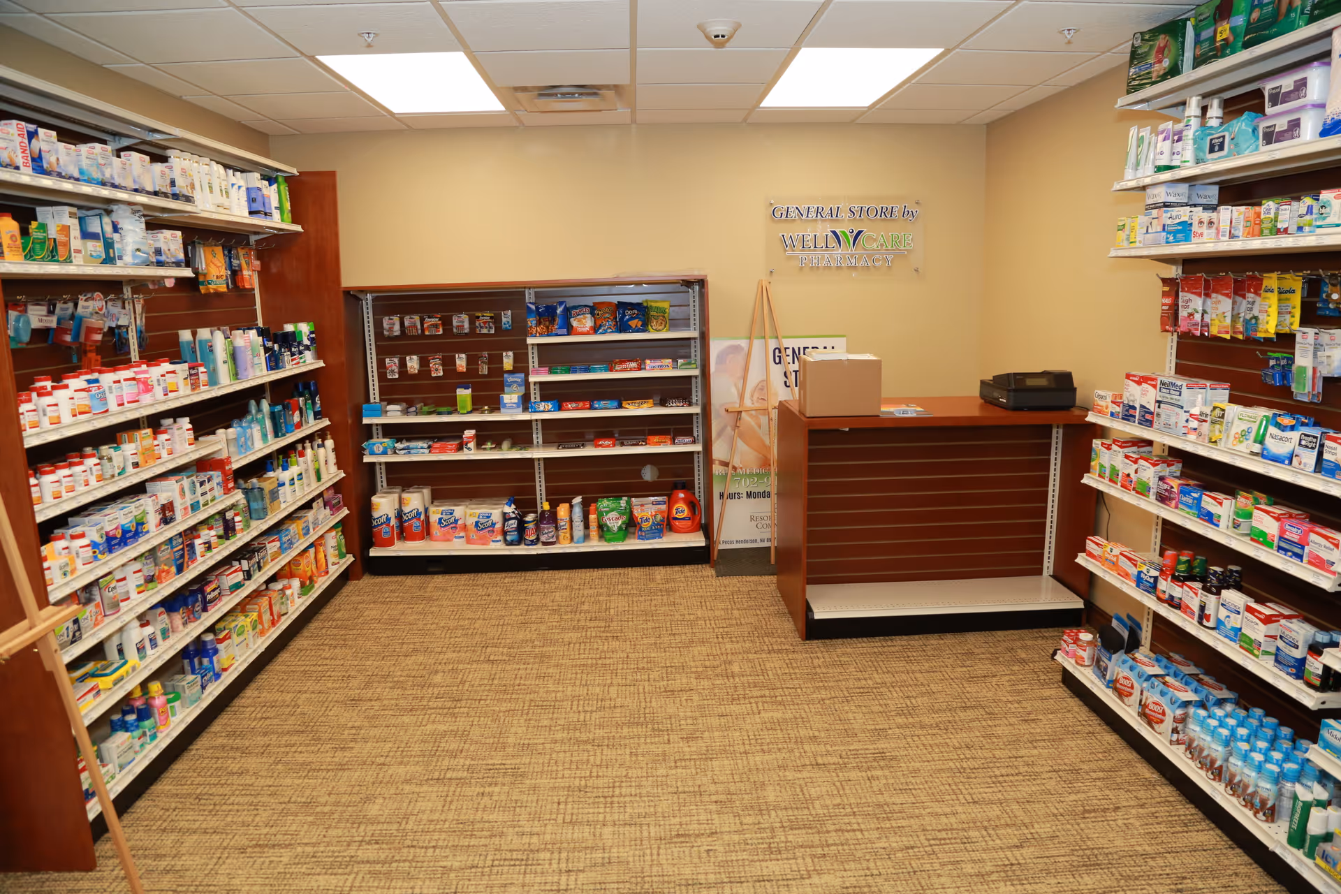 Interior view of a small general store area inside a facility, with shelves stocked with various health and personal care products, snacks, and household items. There is a counter with a cash register and a sign on the wall that reads 'General Store by WellCare Pharmacy'.