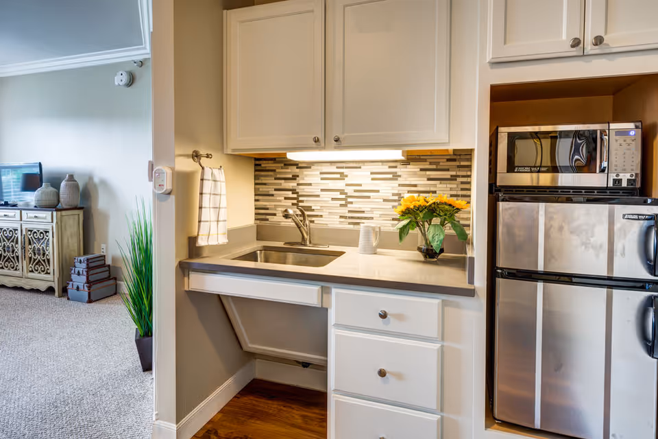 A small kitchen area with a stainless steel sink, a modern faucet, white cabinets, and a tiled backsplash. On the countertop, there is a white mug and a vase with yellow flowers. To the right, there is a stainless steel microwave above a compact refrigerator. In the background, a living room area is partially visible with a TV on a decorative wooden cabinet, some stacked boxes, and a tall green plant.