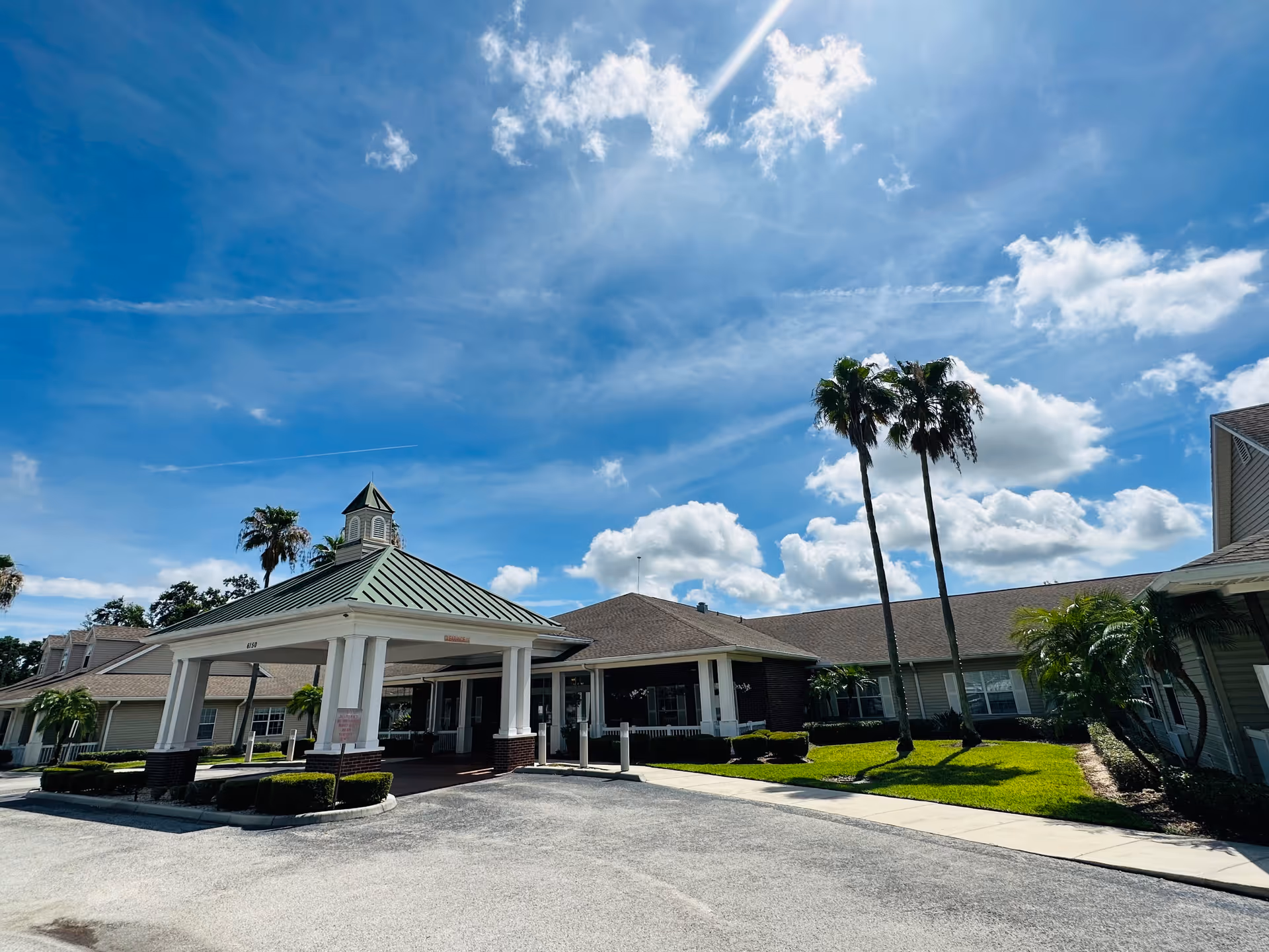 Exterior view of Hawthorne Inn of Lakeland assisted living and memory care facility on a sunny day with blue sky and scattered clouds. The building features a covered entrance with white columns and a green roof, surrounded by palm trees and well-maintained landscaping.