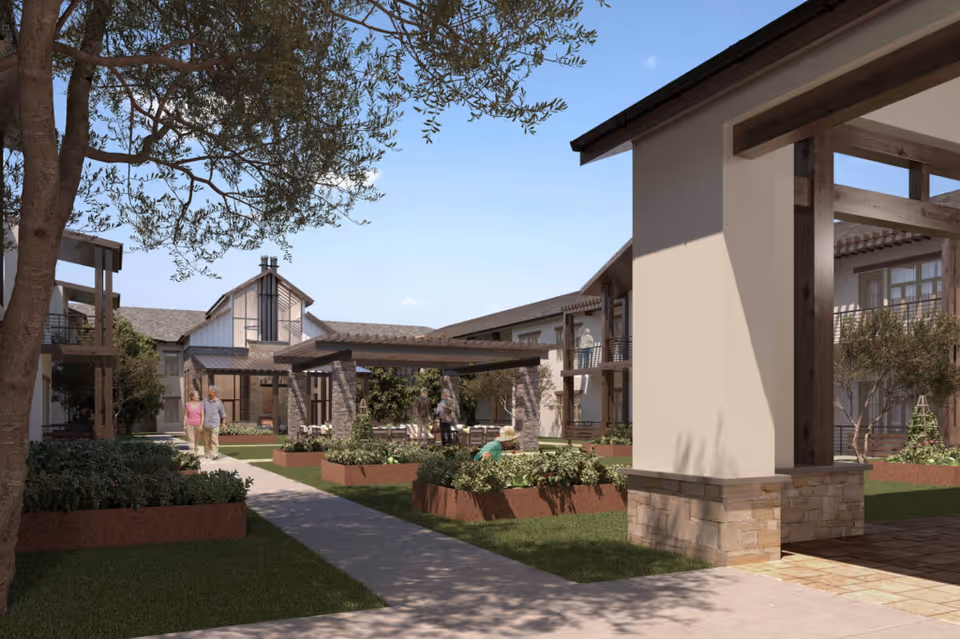 Outdoor courtyard area of a senior living facility with raised garden beds, trees, and a covered seating area. Several people are walking and gardening in the space, surrounded by two-story buildings with balconies.