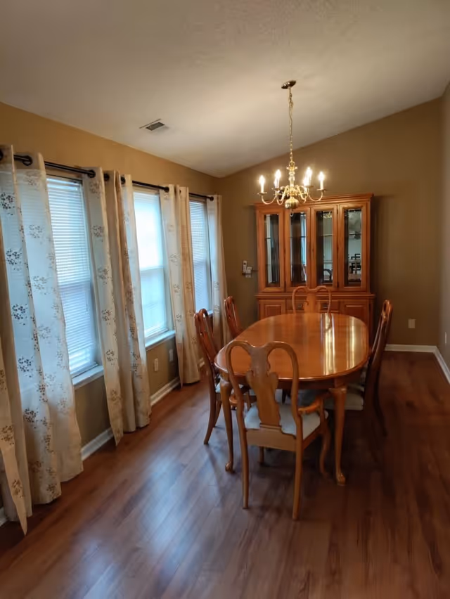 A dining room with a wooden oval dining table surrounded by six matching chairs with cushioned seats. The room has three windows with floral-patterned curtains on the left wall, and a wooden china cabinet with glass doors against the far wall. A brass chandelier with lit bulbs hangs above the table. The floor is wooden, and the walls are painted beige.