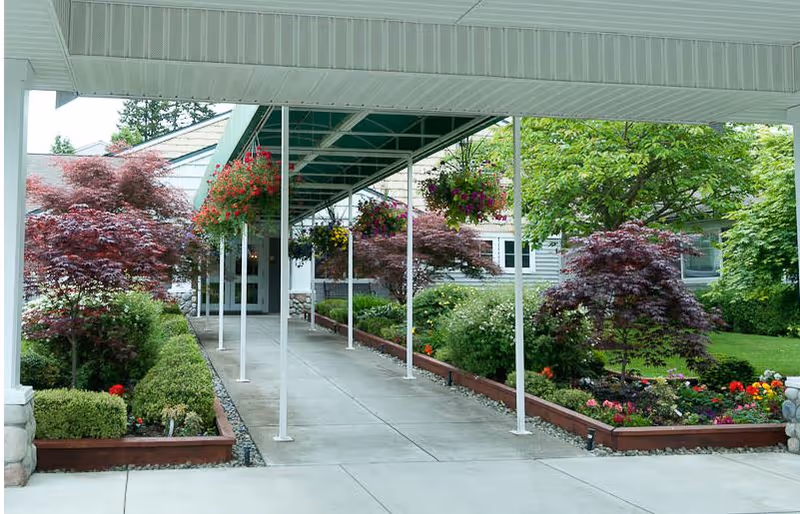Covered walkway leading to the entrance of a building, flanked by landscaped garden beds with various shrubs, small trees, and hanging flower baskets.