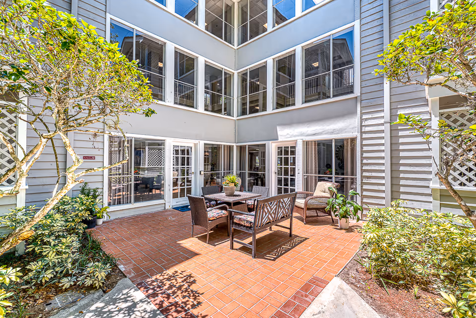 Outdoor courtyard area with brick flooring, surrounded by a multi-story building with large windows and glass doors. The courtyard features a table with four chairs and a bench, along with potted plants and greenery on the sides.