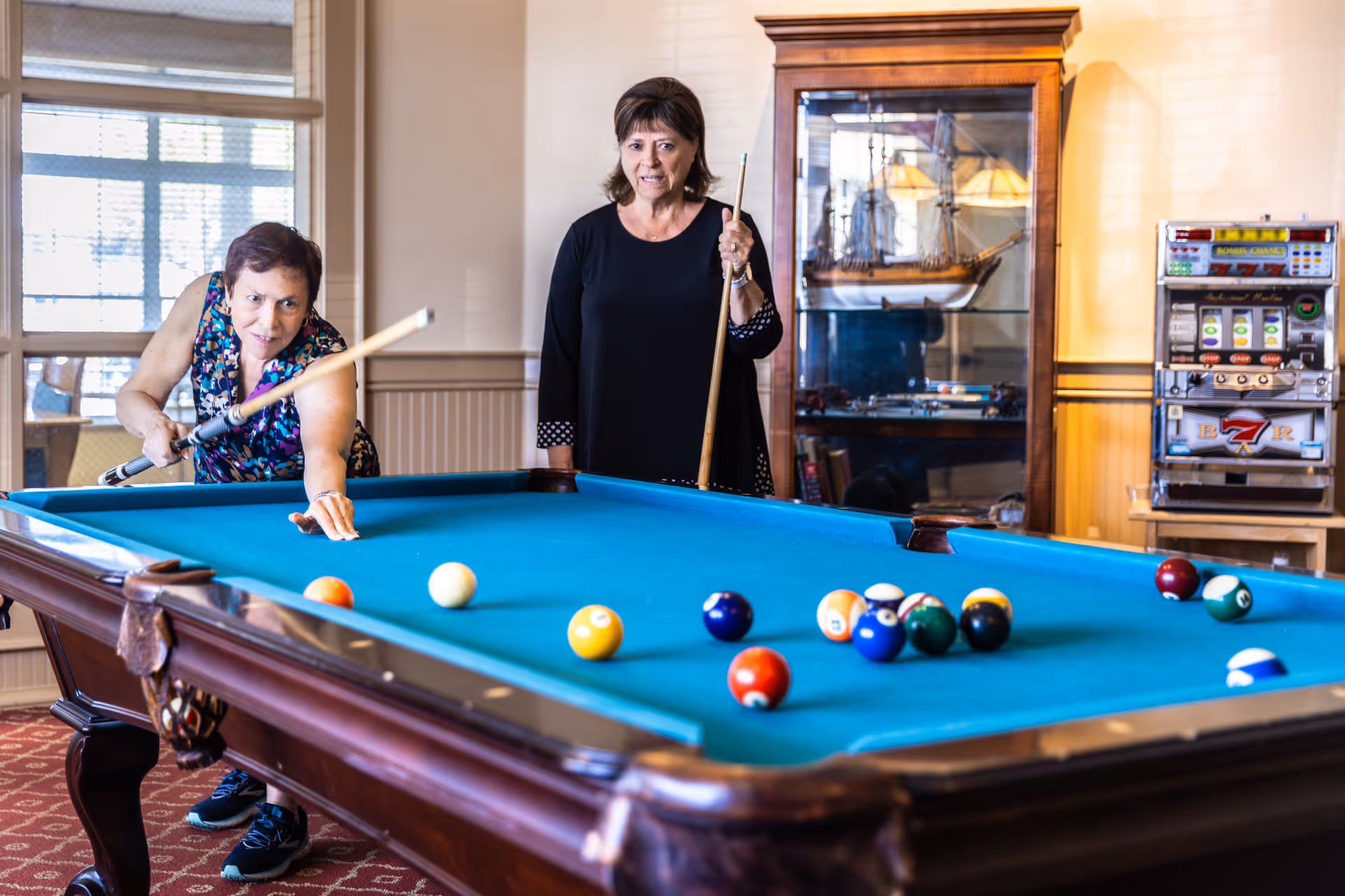 Two women playing pool in a well-lit room with a large window, a wooden display cabinet with a model ship, and a slot machine in the background.
