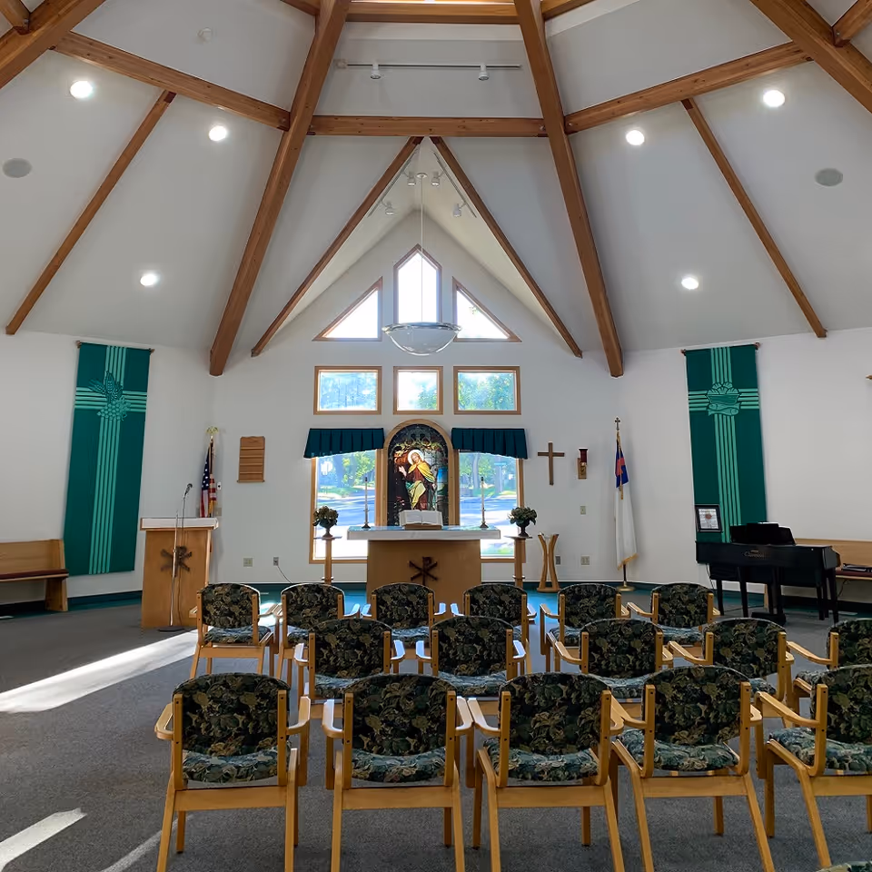 Sloped wooden ceiling beams frame a chapel with altar, banners, and floral-patterned chairs.