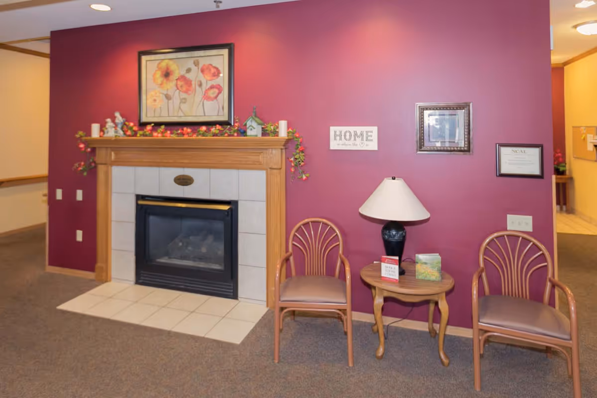 A cozy interior corner featuring a fireplace with a wooden mantel decorated with flowers and small figurines. Above the mantel is a framed floral painting. To the right of the fireplace, there are two wooden chairs with cushions and a small wooden table between them holding a lamp and some books. The wall behind is painted a deep red color and has framed pictures and a sign that reads 'HOME is where the ♥ is'.