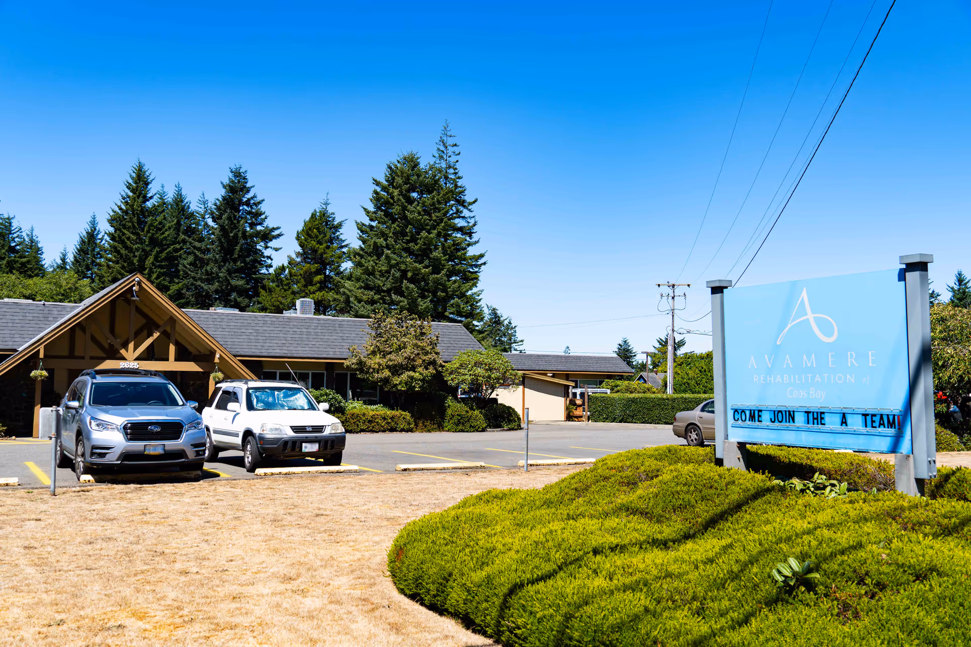 Exterior view of Avamere Rehabilitation of Coos Bay facility with a parking lot containing several cars, a blue sign with the facility name and a message board, green bushes, and tall trees in the background under a clear blue sky.