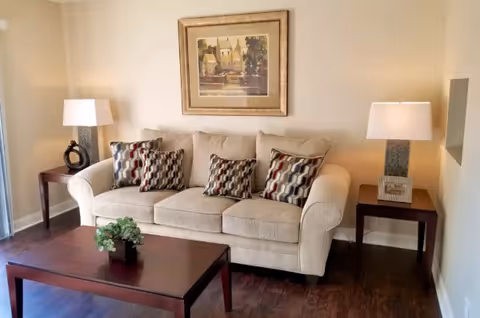 A cozy living room with a beige sofa adorned with four patterned cushions. On either side of the sofa are dark wooden side tables, each holding a lamp with a white shade. Above the sofa hangs a framed painting of a scenic village. In front of the sofa is a dark wooden coffee table with a small green plant. The room has wooden flooring and light-colored walls.