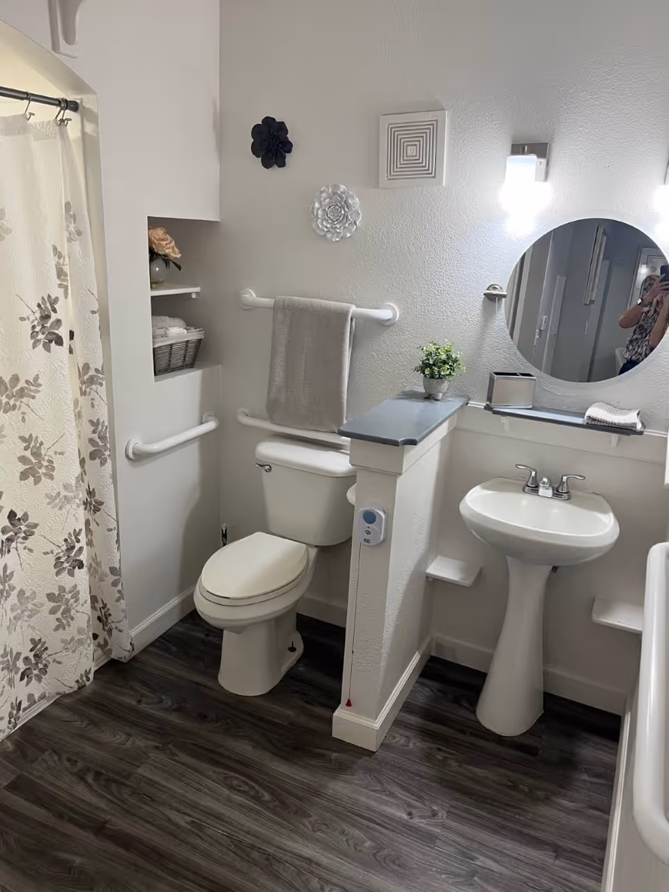 A clean and well-lit bathroom with a white toilet, pedestal sink, and a round mirror above the sink. There is a towel hanging on a grab bar above the toilet, decorative wall hangings, a small potted plant on a half wall, and a floral-patterned shower curtain. The floor has dark wood-like vinyl flooring.