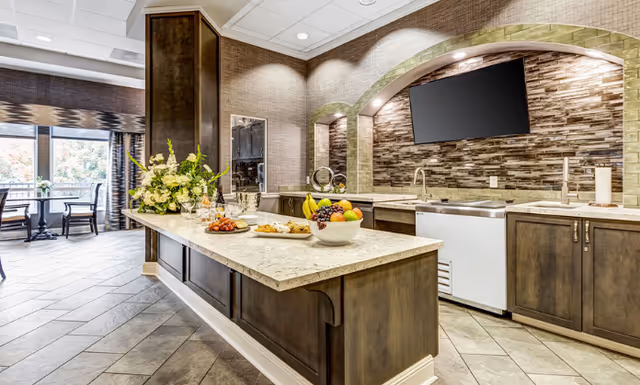 A bright communal kitchen and serving island in a dining area with fruit bowls, flowers, a sink, and a wall-mounted TV.