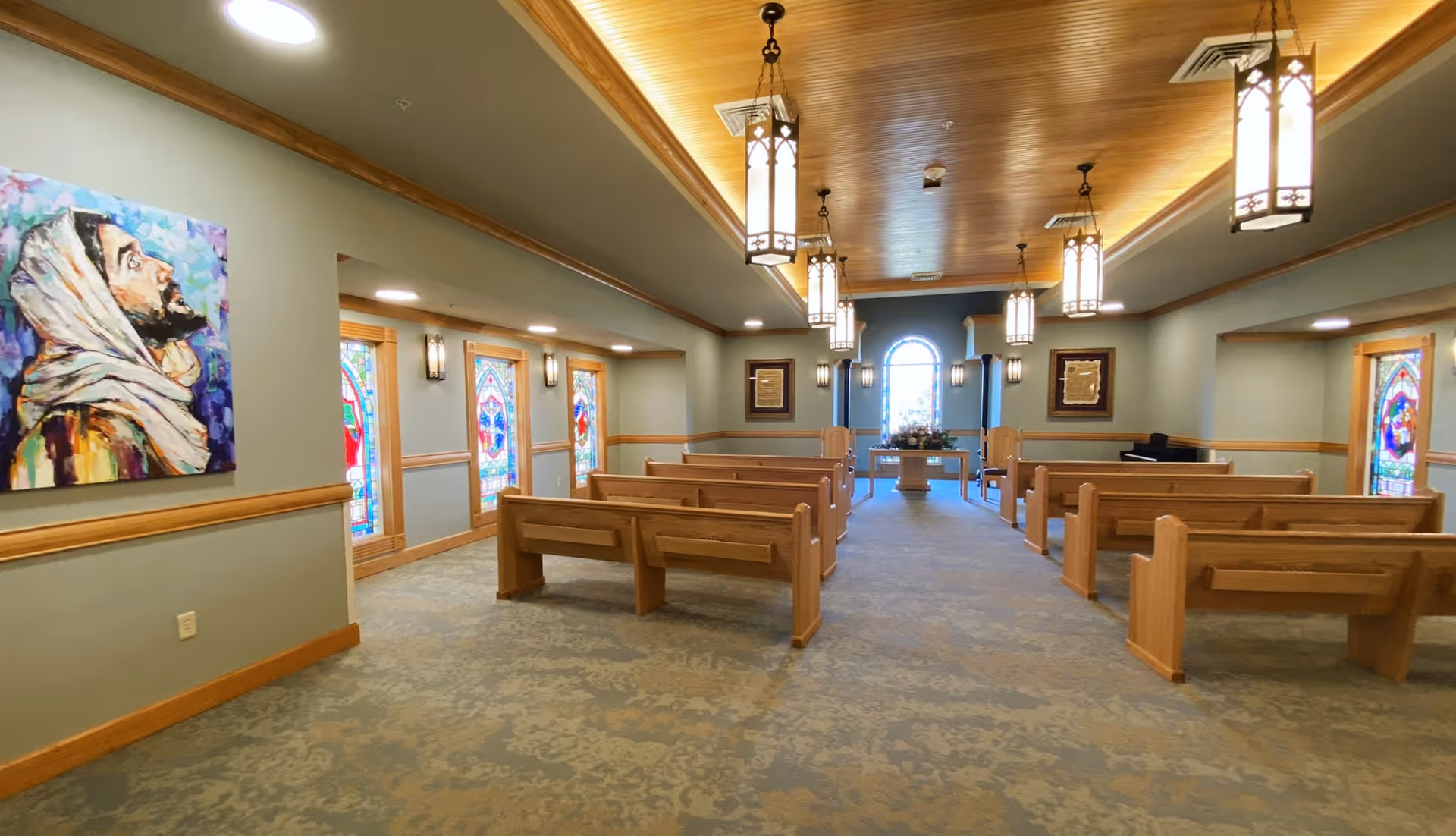 Interior view of a chapel or worship room with wooden pews arranged in rows facing an altar with flowers. The room has stained glass windows along the side walls, a painting of a man with a head covering on the left wall, and hanging lantern-style ceiling lights. The ceiling is wooden with recessed lighting and the walls are painted in a soft green color.