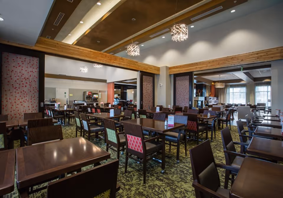 A spacious dining room with multiple wooden tables and chairs, some with colorful patterned backs. The room features a green patterned carpet, modern chandeliers hanging from a ceiling with wooden beams, and large windows allowing natural light to enter. The walls have decorative panels with a floral design.