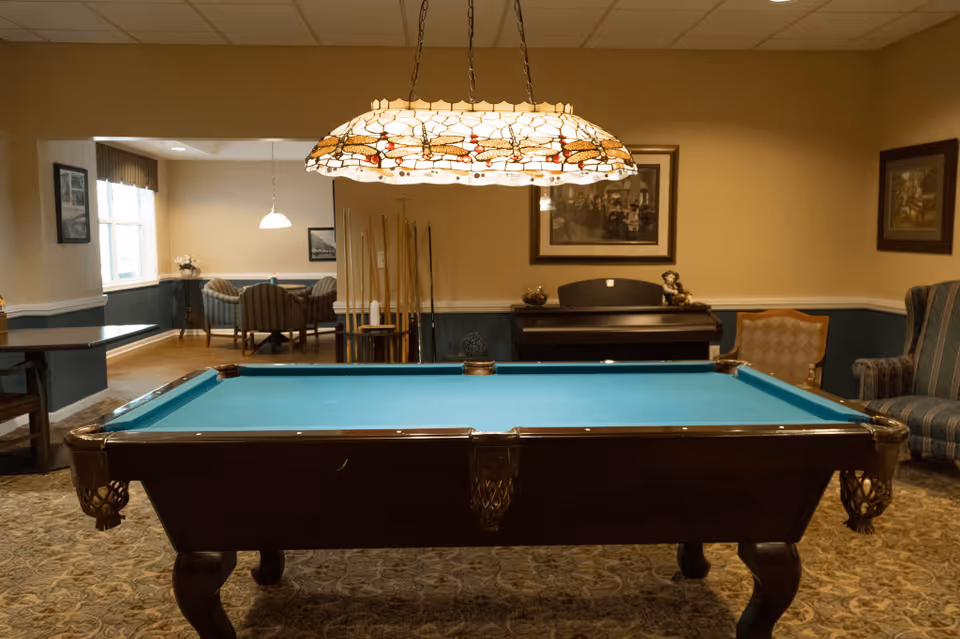 Indoor game room with a billiards table under a stained-glass hanging lamp and seating in the background.