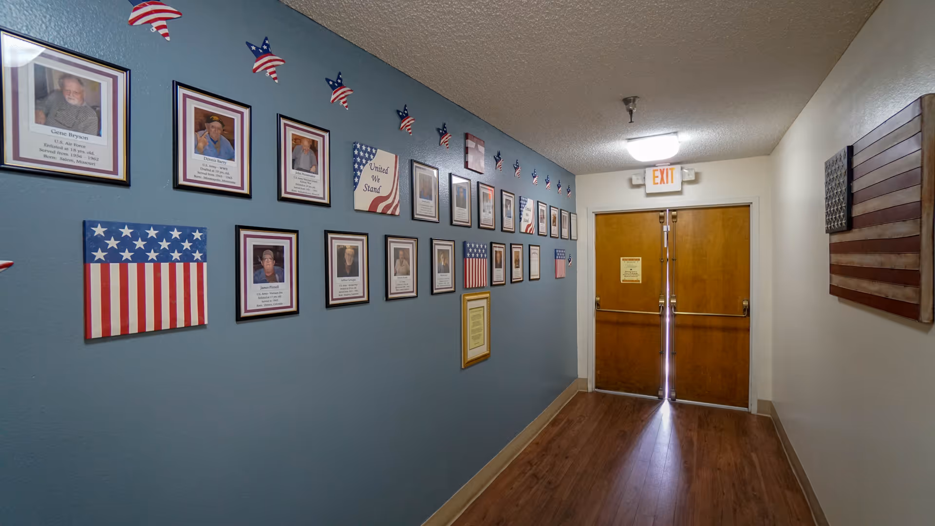 A hallway with wooden double doors at the end, an exit sign above the doors, and a blue wall on the left side decorated with framed photos and patriotic American flag-themed decorations. The right wall is plain with a wooden American flag decoration.