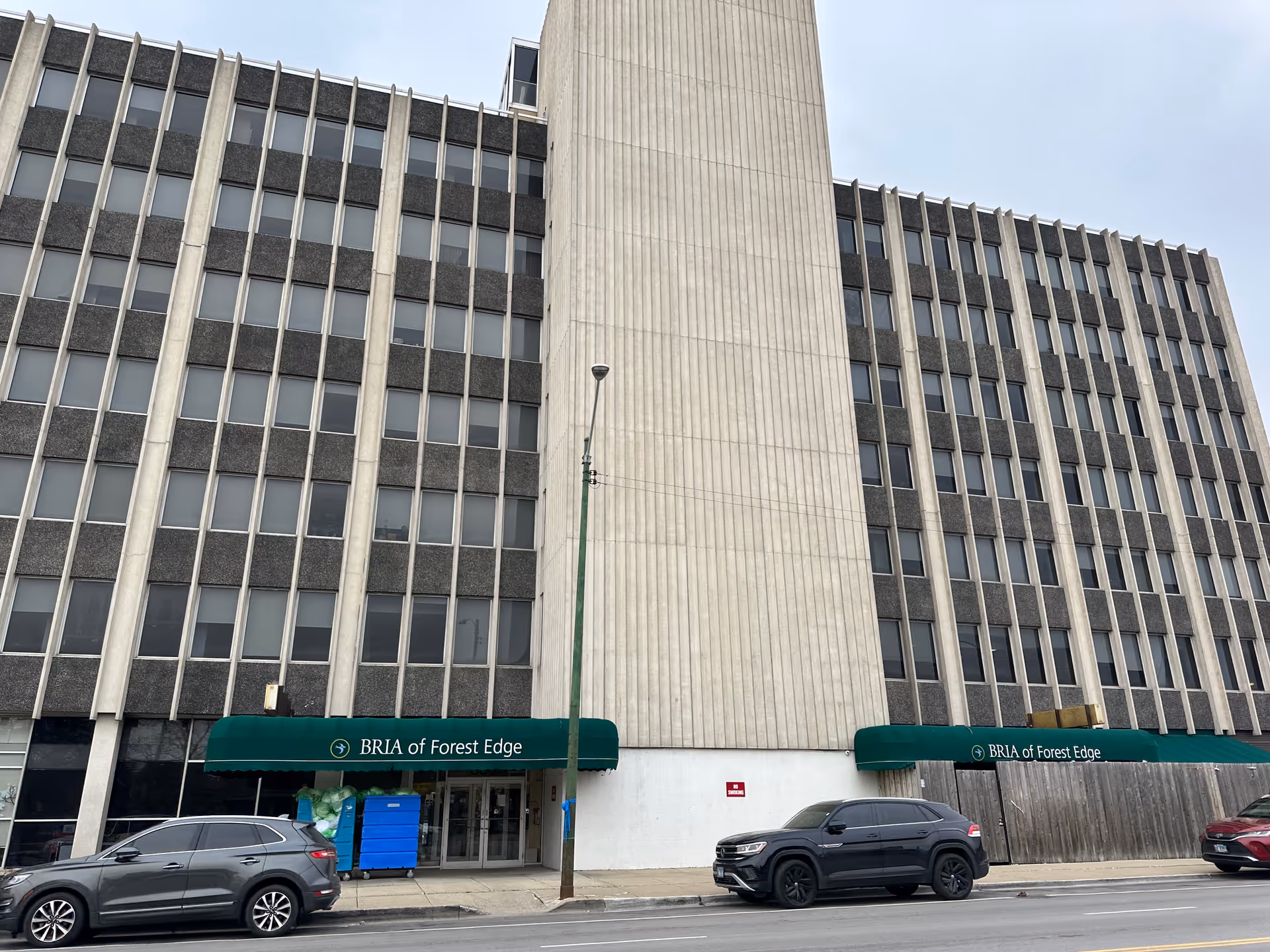 Exterior view of a multi-story building with green awnings labeled 'BRIA of Forest Edge' above the entrances. Several cars are parked along the street in front of the building, and a streetlamp is visible in the center.