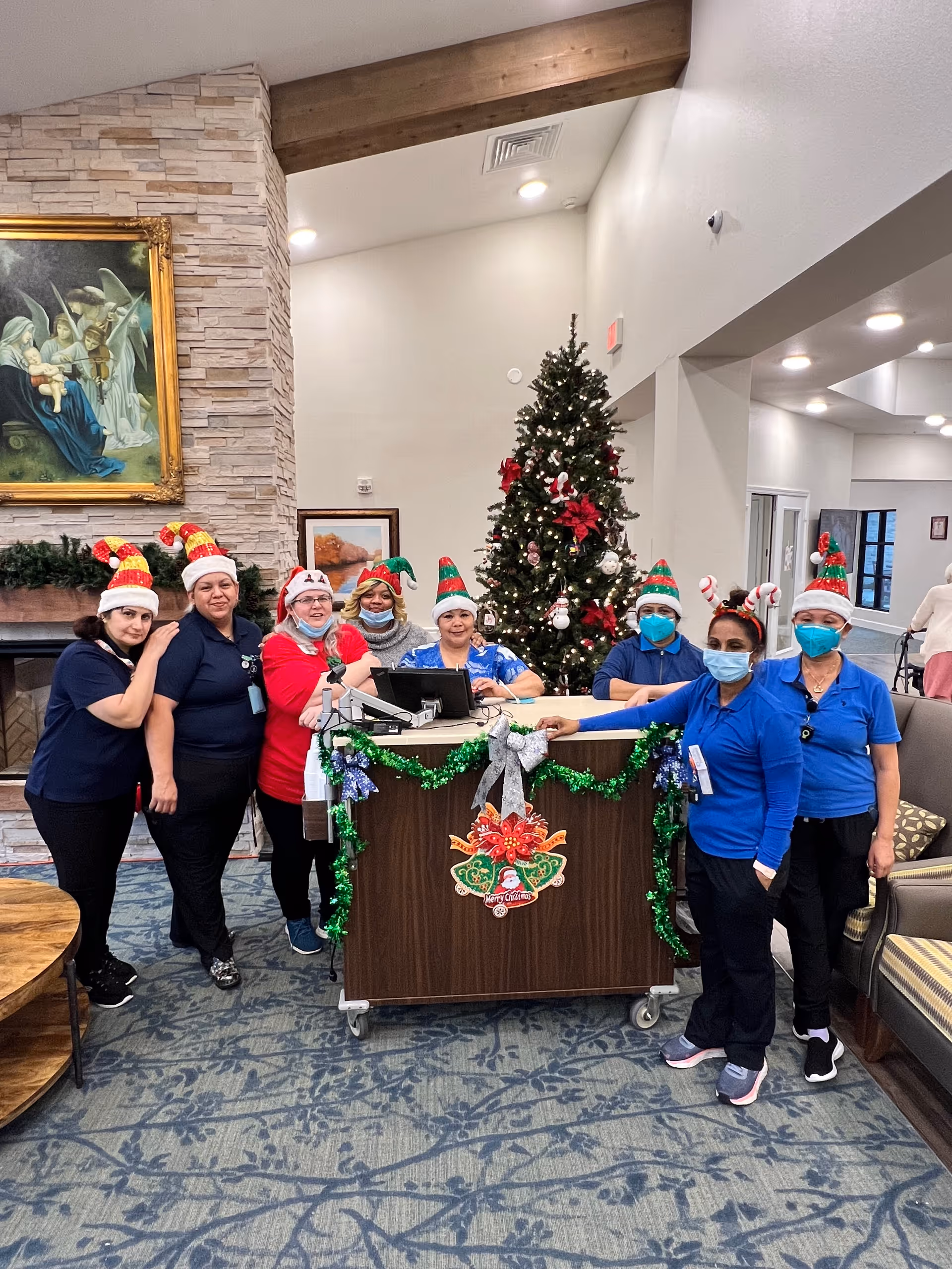 A group of seven staff members wearing festive holiday hats and face masks standing around a decorated reception desk in a senior living facility lobby. Behind them is a decorated Christmas tree and a stone fireplace with a framed painting above it. The room has high ceilings with wooden beams and recessed lighting.