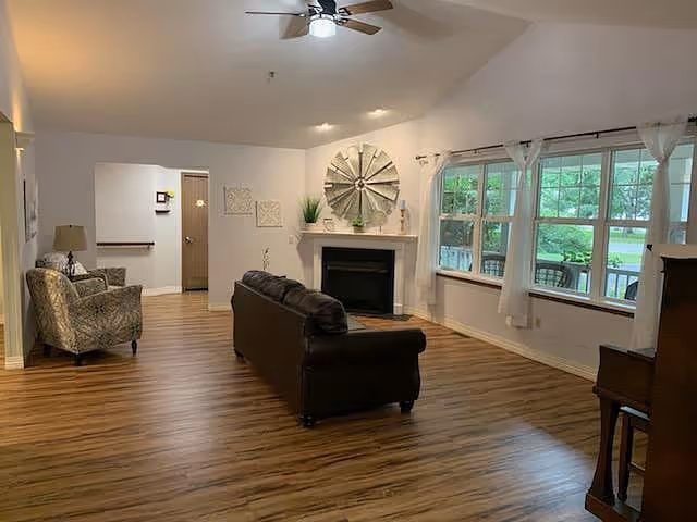 A spacious living room with wooden flooring, a dark brown leather sofa facing a white fireplace with decorative items above it, a patterned armchair with a lamp on a side table, large windows with white curtains letting in natural light, and a ceiling fan with lights.