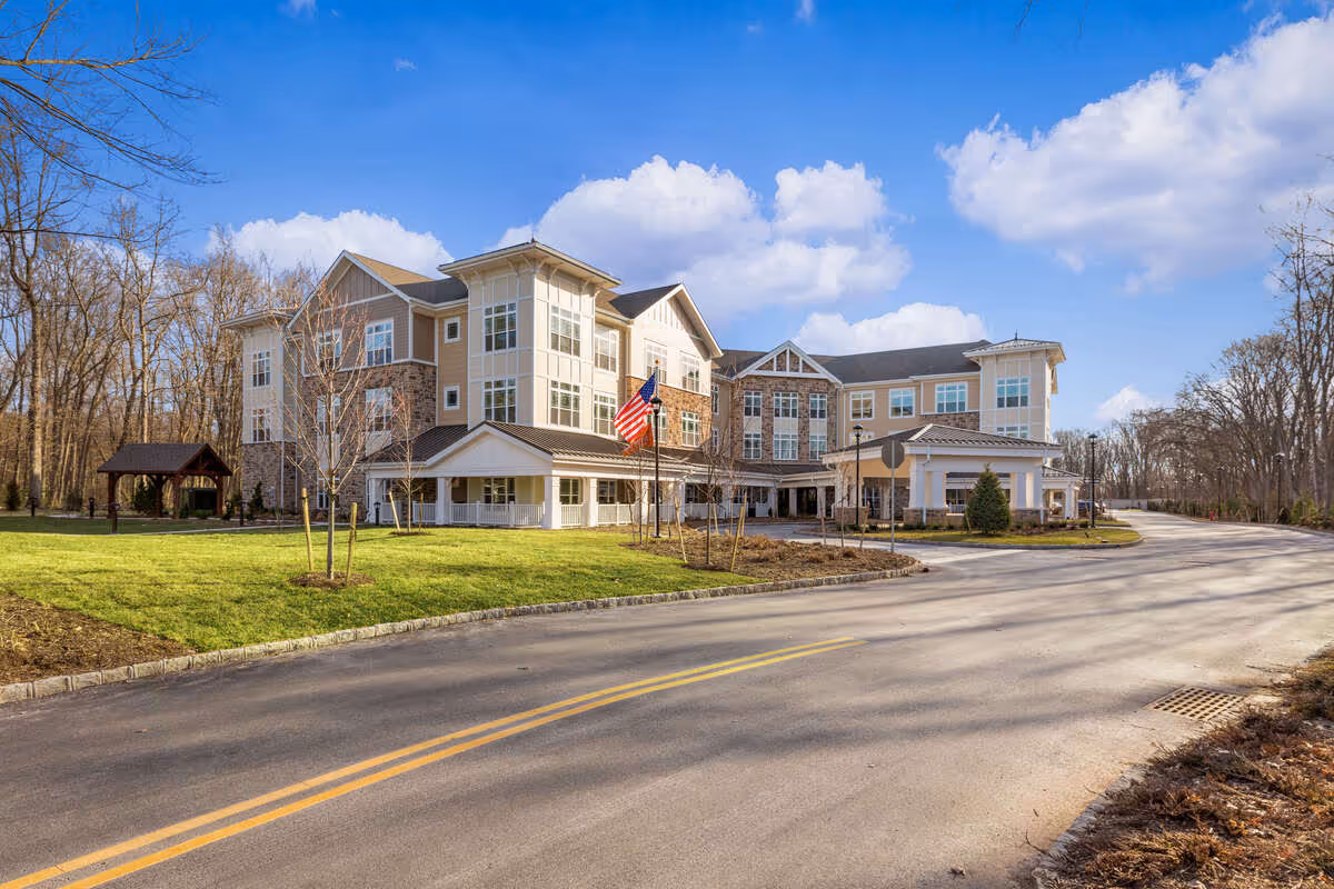 Large three-story senior living building with an American flag by the covered entrance, a curved driveway, and grassy landscaping under a blue sky.