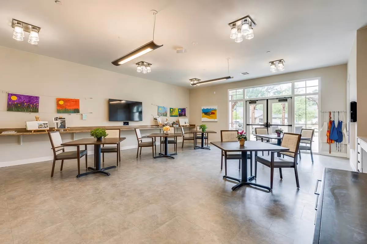Bright communal activity room with several small tables and chairs, a wall-mounted TV, artwork on the walls, and large windows and doors letting in natural light.