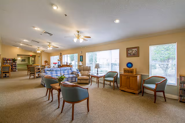 A spacious and well-lit common area in a senior living facility featuring multiple seating options including a plaid sofa, armchairs, and wooden chairs with cushions. Large windows allow natural light to fill the room, and ceiling fans provide ventilation. There are small tables with lamps and decorative items, a bookshelf, and a cabinet with a decorative plate on top.