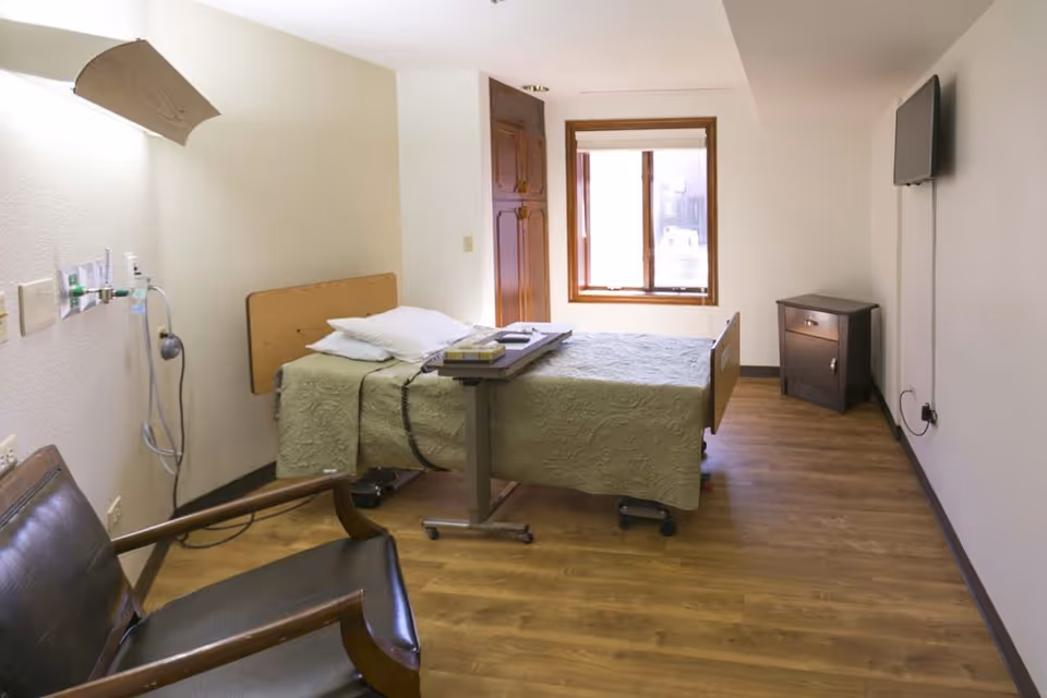 A simple, clean patient room with a single bed covered in a green quilt, a small overbed table with books, a wooden chair with black cushioning, a window with blinds, a wall-mounted TV, and a small wooden cabinet. The room has wood flooring and white walls.