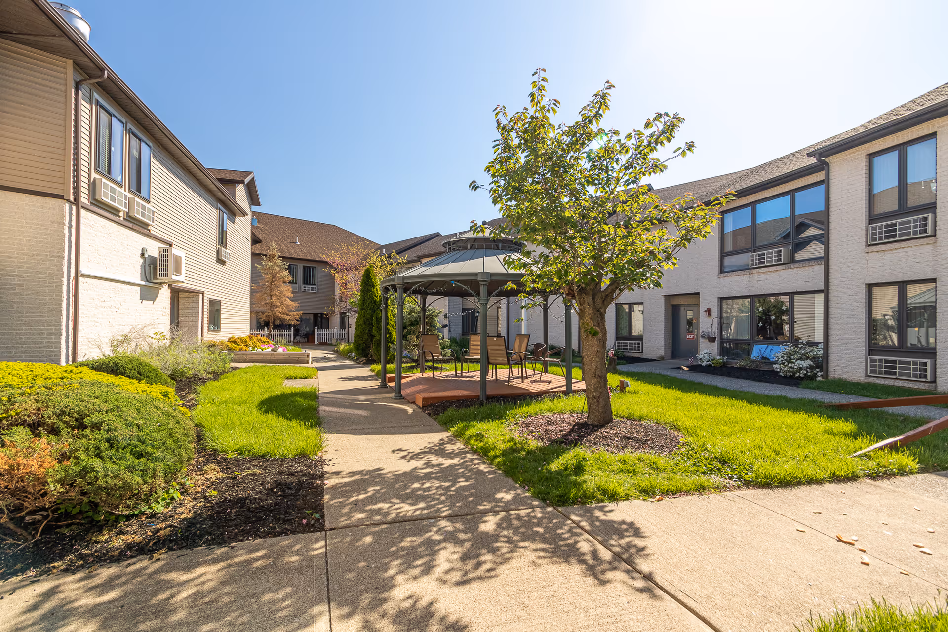 Outdoor courtyard area of a senior living facility with a paved walkway, green grass, bushes, a tree, and a gazebo with chairs underneath. Surrounding the courtyard are two-story buildings with multiple windows and air conditioning units.