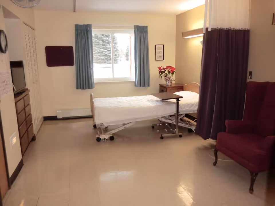 A simple patient room in a senior living facility with a hospital bed, a small overbed table, a wooden nightstand with a poinsettia plant, a maroon armchair, a window with blue curtains, and a privacy curtain partially drawn.