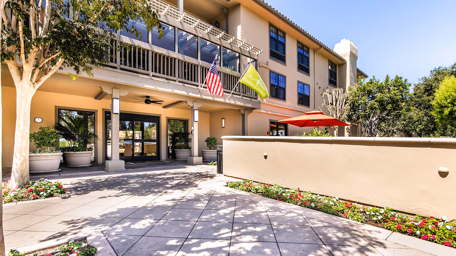Exterior view of Atria San Juan senior living facility showing the entrance area with large glass doors, potted plants, an American flag, a green flag, and a patio with a red umbrella. The building has multiple windows and a balcony above the entrance.