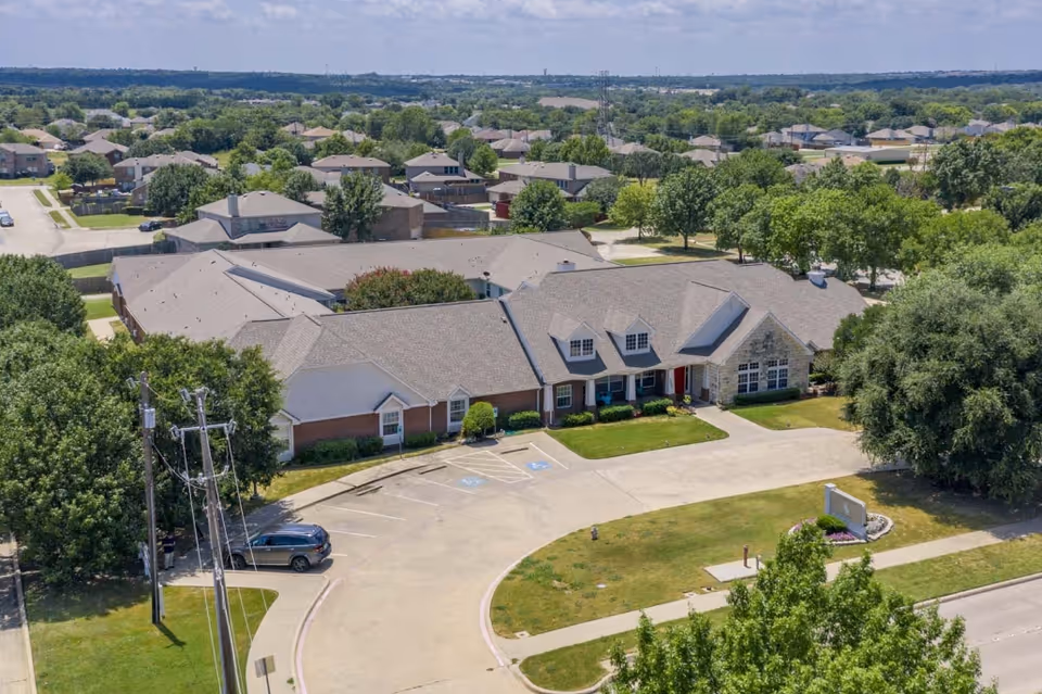 Aerial view of Meadow Creek Senior Living facility showing a single-story building with a large parking area, surrounded by trees and residential houses under a partly cloudy sky.