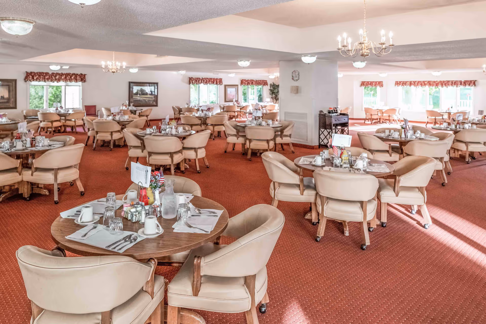 Spacious dining room with multiple round tables set for meals and beige upholstered chairs on a red carpet.